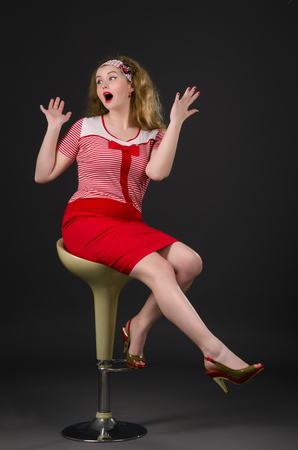 Beautiful young pin-up woman with wow face sitting on the bar chair. Excited girl wearing red cloths with radiant red lipstick. Dark background.の写真素材