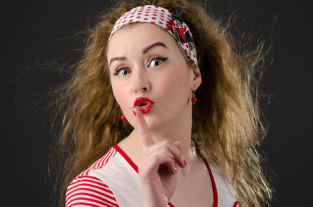 Beautiful young pin-up woman asking for silence. Girl wearing red cloths with radiant red lipstick. Dark background.の写真素材