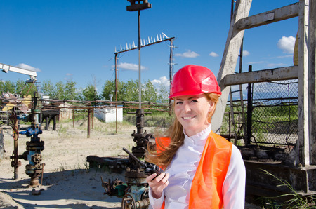 Beautiful woman engineer near well head in the oil field talking on the radio wearing red helmet and work clothes. Pump jack and pipeline background. Oil and gas concept.の写真素材