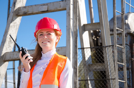 Beautiful woman engineer in the oil field talking on the radio wearing red helmet and work clothes. Pump jack background. Oil and gas concept.の写真素材