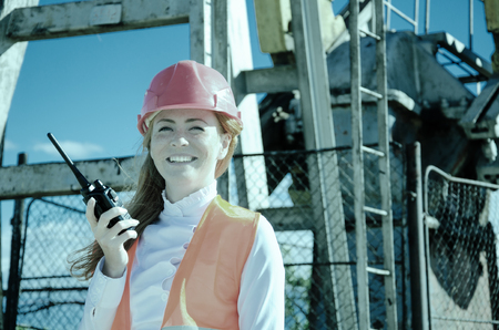 Beautiful woman engineer in the oil field talking on the radio wearing red helmet and work clothes. Pump jack background. Oil and gas concept. Toned.の写真素材