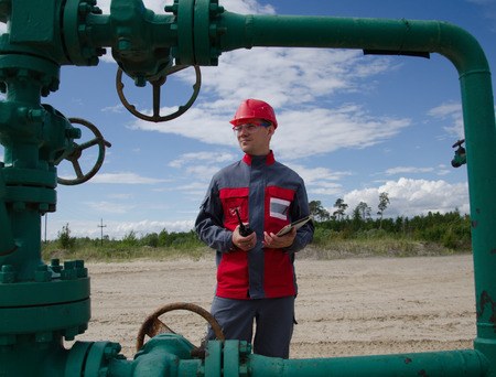 Worker near well head valve holding tablet computer and radio and wearing red helmet in the...