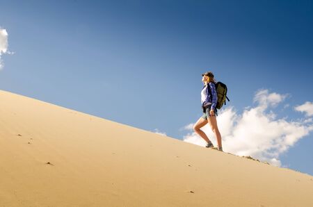 Young beautiful woman backpacker traveling in the desert. Sandy dunes and blue sky on sunny summer day. Travel, adventure, freedom concept. Toned.の写真素材