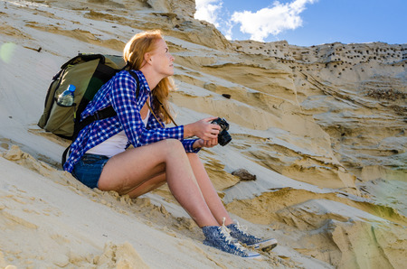 Young beautiful woman backpacker travelling in the desert and making pictures with vintage film camera. Sandy dunes and blue sky on sunny summer day. Travel, adventure, freedom concept. Toned.の写真素材