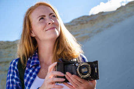 Young beautiful woman backpacker traveling in the desert and making pictures with vintage film camera. Sandy dunes and blue sky on sunny summer day. Travel, adventure, freedom concept. Toned.の写真素材