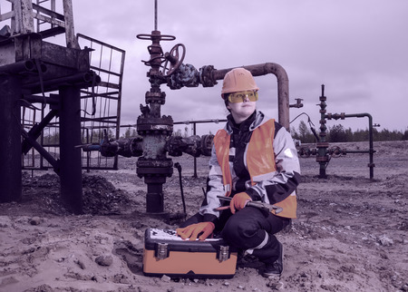 Woman engineer in the oil field with the wrench and toolbox wearing orange helmet and work clothes. Pump jack and wellhead background. Oil and gas concept. Toned.の写真素材