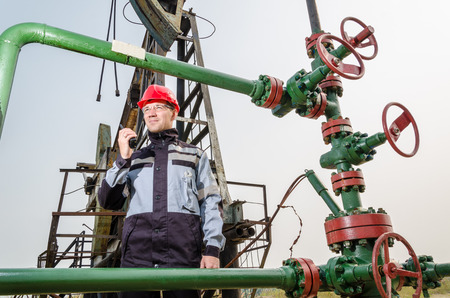 Oilfield worker near wellhead wearing red helmet and work clothes, holding the radio. Pump jack background. Oil and gas concept.の写真素材