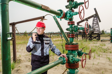 Oilfield worker near wellhead valve, wearing red helmet and work clothes talking on the radio. Pump jack background. Oil and gas concept.の写真素材