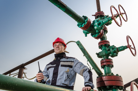 Oilfield worker near wellhead valve, wearing red helmet and work clothes talking on the radio. Oil and gas concept.の写真素材