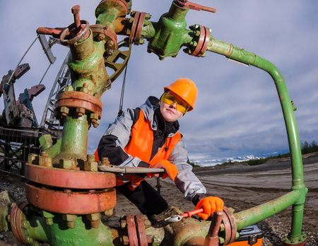 Woman engineer in the oilfield repairing wellhead with the wrench wearing orange helmet and work clothes. Oil rig background. Oil and gas concept. Fish eye shot.の写真素材