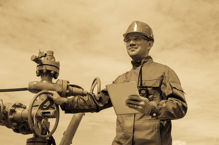 Worker near well head valve holding tablet computer and wearing helmet in the oilfield. Oil and gas concept. Toned.の写真素材