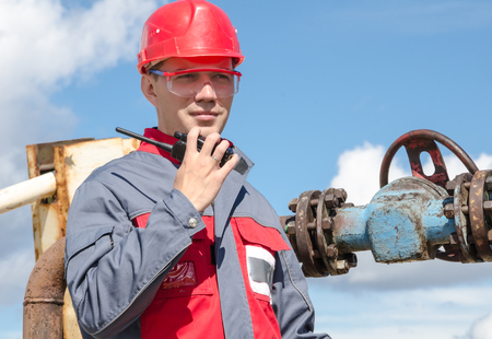 Worker near wellhead valve holding radio and wearing red helmet in the oilfield. Oil and gas concept.の写真素材