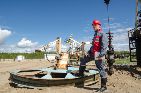 Worker near broken pump jack holding radio and tablet computer in the oilfield. Pump jacks and wellhead background. Oil and gas concept.の写真素材