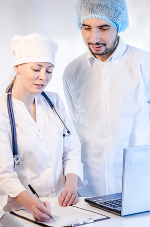 Group portrait of medical doctors in hospital looking at the papers and laptop. Man wearing surgeon coat and woman wearing white cap.の写真素材
