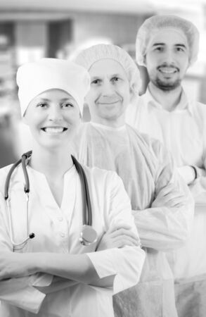 Group portrait of mixed aged medical doctors standing in hospital, looking at camera and smiling. Two men wearing surgeon coat and woman wearing white cap and coat. Focus on woman doctor. Black and white.の写真素材