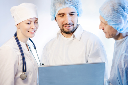 Group portrait of medical doctors in hospital looking at the laptop. Two men wearing surgeon coat and woman wearing white cap. Focus on man doctor.の写真素材