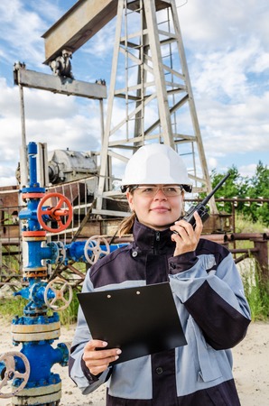 Woman engineer in the oilfield talking on the radio wearing white helmet and work clothes. Industrial site background. Oil and gas concept.の写真素材
