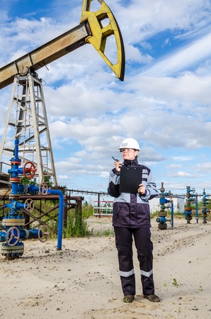 Woman engineer in the oilfield talking on the radio wearing white helmet and work clothes. Industrial site background. Oil and gas concept.の写真素材