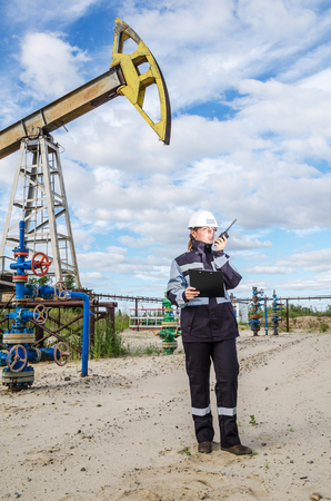 Woman engineer in the oilfield talking on the radio wearing white helmet and work clothes. Industrial site background. Oil and gas concept.の写真素材