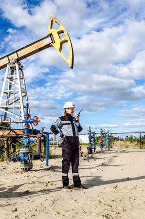 Woman engineer in the oilfield talking on the radio wearing white helmet and work clothes. Industrial site background. Oil and gas concept.の写真素材