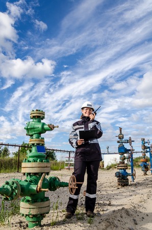 Woman engineer in the oilfield talking on the radio wearing white helmet and work clothes. Industrial site background. Oil and gas concept.の写真素材
