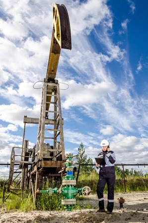 Woman engineer in the oilfield near pump jack wearing white helmet and work clothes. Industrial site background. Oil and gas concept.の写真素材