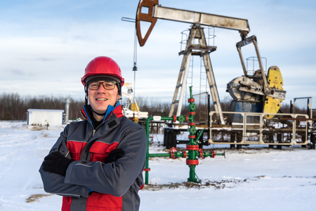 Man worker in the oilfield near pump jack and wellhead, wearing helmet and work clothes. Industrial site background. Oil and gas concept.の写真素材