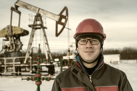 Man worker in the oilfield near pump jack and wellhead, wearing helmet and work clothes. Industrial site background. Oil and gas concept.の写真素材