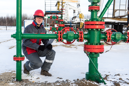 Man worker in the oilfield near pump jack and wellhead, wearing helmet and work clothes. Industrial site background. Oil and gas concept.の写真素材