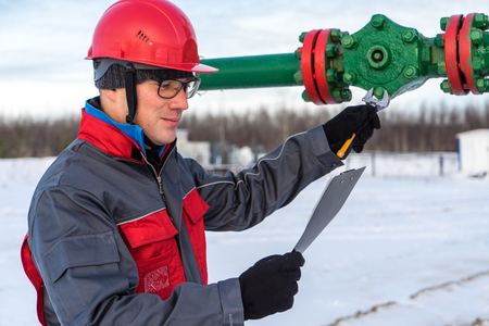Man worker in the oilfield near pump jack and wellhead, wearing helmet and work clothes. Industrial site background. Oil and gas concept.の写真素材