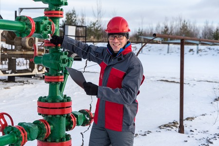 Man worker in the oilfield near pump jack and wellhead, wearing helmet and work clothes. Industrial site background. Oil and gas concept.の写真素材