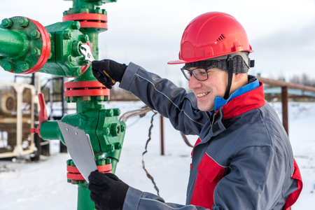 Man worker in the oilfield near pump jack and wellhead, wearing helmet and work clothes. Industrial site background. Oil and gas concept.の写真素材