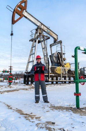 Man worker in the oilfield near pump jack and wellhead, wearing helmet and work clothes. Industrial site background. Oil and gas concept.の写真素材