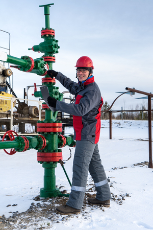 Man worker in the oilfield near pump jack and wellhead, wearing helmet and work clothes. Industrial site background. Oil and gas concept.の写真素材