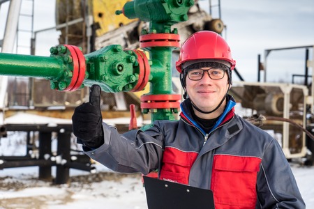 Man worker in the oilfield near pump jack and wellhead, wearing helmet and work clothes. Industrial site background. Oil and gas concept.の写真素材