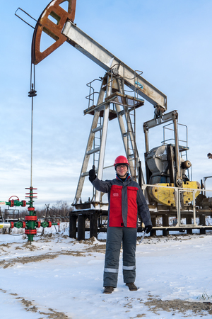 Man worker in the oilfield near pump jack and wellhead, wearing helmet and work clothes. Industrial site background. Oil and gas concept.の写真素材