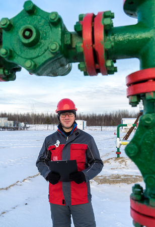 Man worker in the oilfield near pump jack and wellhead, wearing helmet and work clothes. Industrial site background. Oil and gas concept.の写真素材