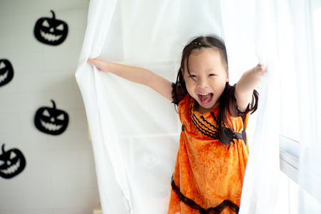 Portrait Asian little girl playing and hiding in the white clean curtain with Halloween decoration in background.の写真素材