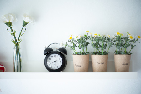 Vintage alarm clock puts on the white wooden shelf decorated by the blossom flowers in the pottery with blurred background of vaseの写真素材