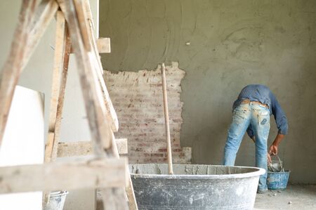 Selective focus on wooden holder in the mixed cement bucket with blurred worker and plastered cement on bricklayer wall in background at the construction siteの写真素材
