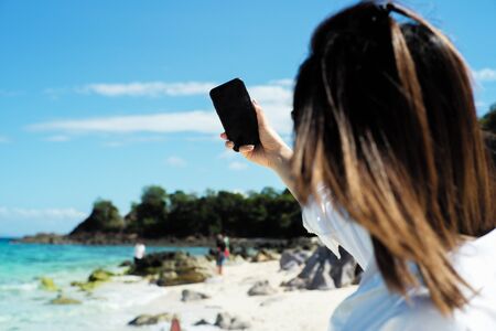 Back portrait of female tourist holds mobile phone and takes selfie picture with blurred seascape and people in background. Selective focus on blank screen of mobile held by woman for selfieの写真素材