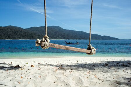Selective focus on the old wooden swing hanging from the tree with defocused traditional long tail boat sailing in the sea in background.の写真素材