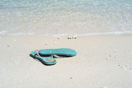 Closeup pair of old sandals put on the white sand beach with blurred sea surfing into the coast backgroundの写真素材