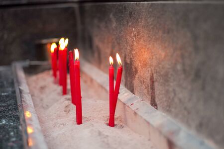 Group of red lighting candles put in the sand in concrete tray with blurred hands holding incensesの写真素材
