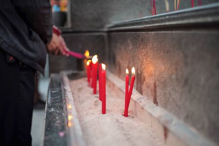 Group of red lighting candles put in the sand in concrete tray with blurred hands holding incensesの写真素材