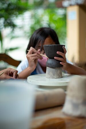Closeup hands of little girl molding the clay on the wheel tray with blurred portrait of girl with facial mask in backgroundの写真素材