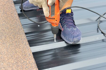 Closeup hands of workers installing the metal sheet roof by electrical drilling machine. Selective focus on the drilling tool while building the roofの写真素材