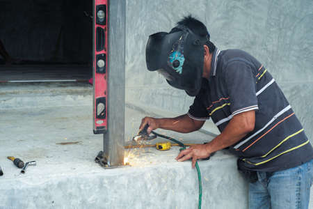 Welder with face shield is welding the steel column with the plate on the concrete floor at the construction siteの写真素材