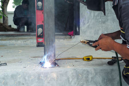 Welder with face shield is welding the steel column with the plate on the concrete floor at the construction siteの写真素材