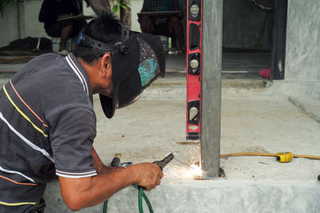 Welder with face shield is welding the steel column with the plate on the concrete floor at the construction siteの写真素材